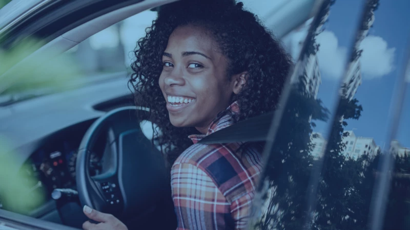 A woman wearing a colorful plaid shirt grips the steering wheel and smiles through the open window of a vehicle.