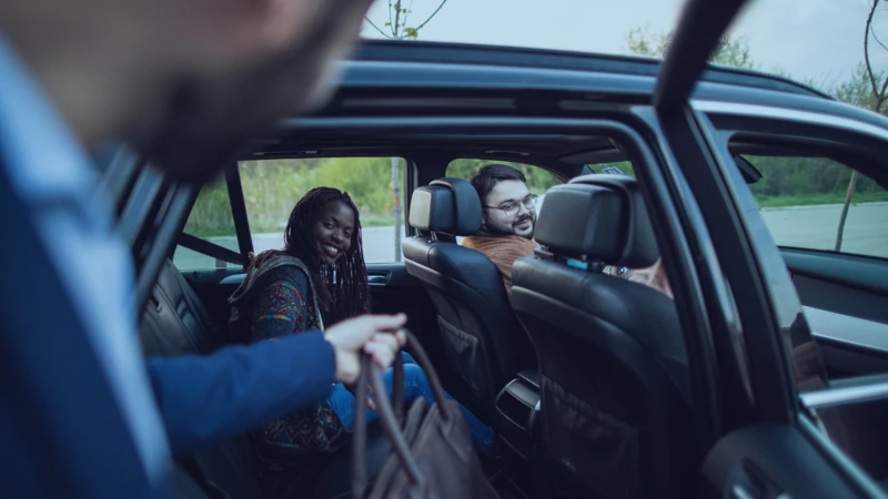 Man handing a suitcase into a rideshare vehicle with driver and passenger smiling